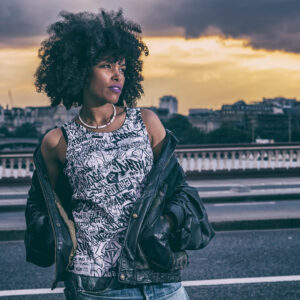 GOLOKA Women category tile: model with an afro wearing a graffiti print tank top and leather jacket on a bridge at sunset